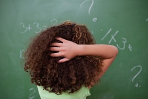 Schoolgirl thinking about algebra while scratching the back of her head in front of a blackboard