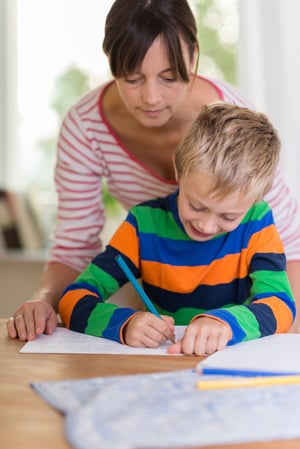 Teacher or concerned mother looking over the shoulder of a young boy as he sits at a table writing or drawing on a sheet of paper